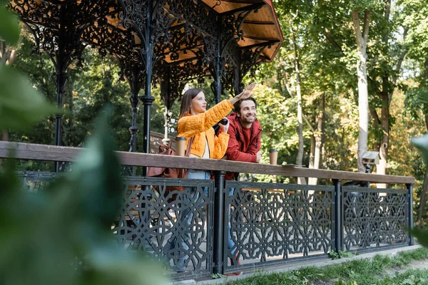 Young woman holding binoculars and pointing with hand near boyfriend and coffee to go on railing outdoors — Stock Photo