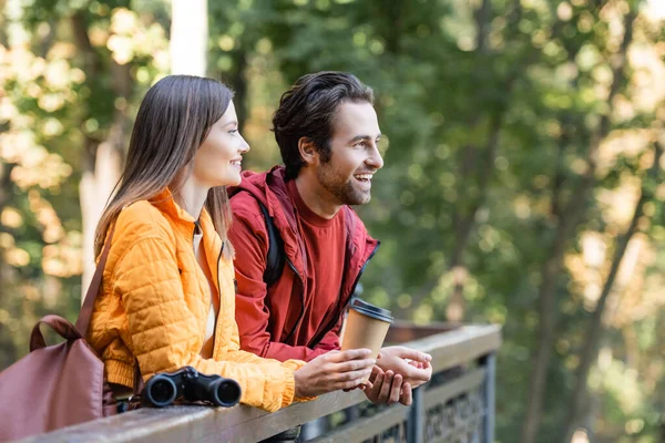 Positive woman with backpack holding paper cup near boyfriend and binoculars on railing outdoors — Stock Photo