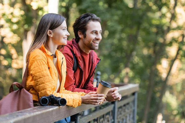 Voyageur souriant tenant à emporter boisson près du petit ami et jumelles sur rambarde à l'extérieur — Photo de stock