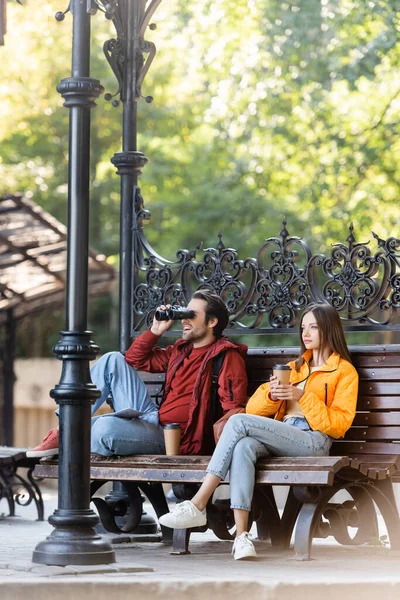 Turista feliz mirando a través de prismáticos cerca de novia con café para ir sentado en el banco al aire libre - foto de stock