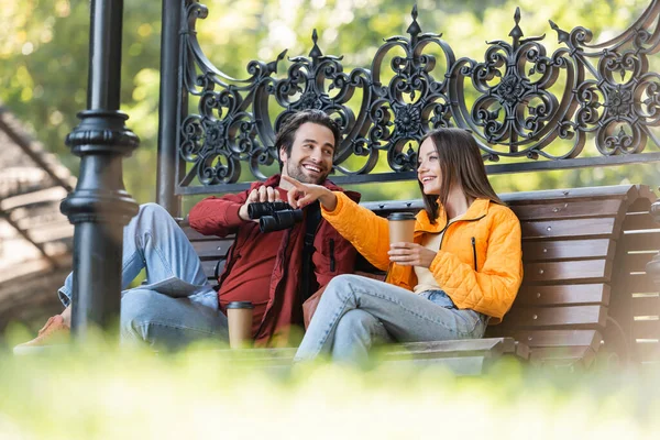 Viajero sonriente sosteniendo café y señalando con el dedo cerca de novio con prismáticos en el banco al aire libre - foto de stock