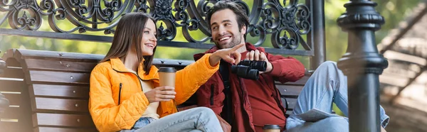 Turista sonriente sosteniendo prismáticos cerca de su novia con taza de papel apuntando con el dedo al aire libre, pancarta - foto de stock