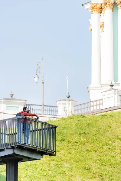 Smiling tourist pointing with finger near girlfriend with binoculars on viewpoint in city — Stock Photo