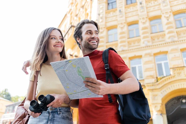 Low angle view of positive tourists with map and binoculars looking away on urban street — Stock Photo