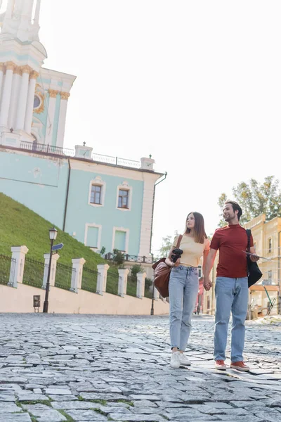 Positive tourist with map and backpack holding hand of girlfriend on urban street — Stock Photo