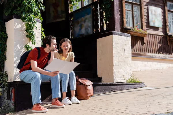 Pensive tourist holding map near girlfriend and building outdoors — Stock Photo