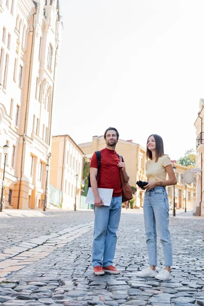 Donna felice in possesso di binocolo vicino fidanzato con mappa e zaini sulla strada della città — Foto stock