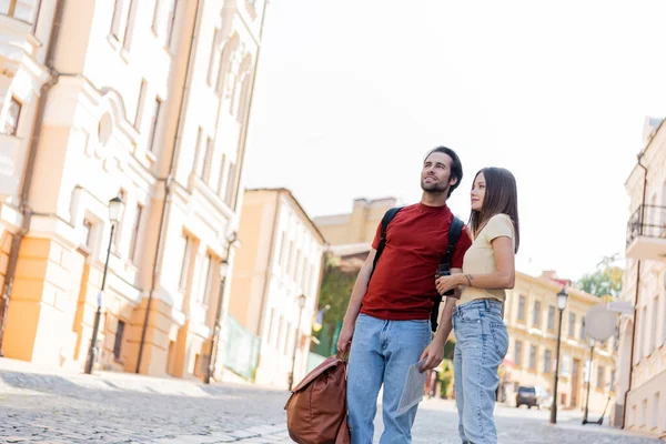 Позитивний мандрівник з картою і рюкзаками, що стоять поруч з подругою, що тримає бінокль на міській вулиці — стокове фото