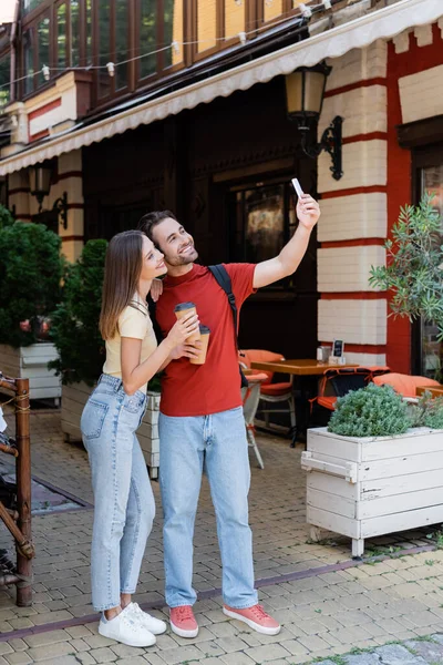 Pareja positiva con mochilas y café para ir a tomar selfie en la calle urbana - foto de stock
