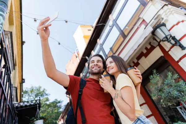 Vista a basso angolo di turisti positivi con tazze di carta che scattano selfie sulla strada urbana — Foto stock