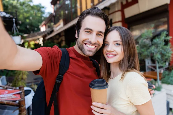 Viajeros positivos con mochila y café para ir mirando a la cámara al aire libre - foto de stock