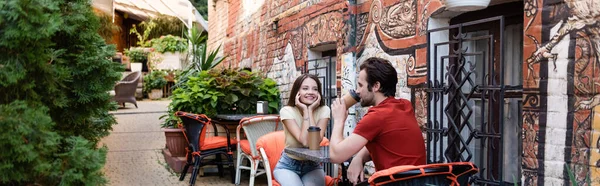 Позитивна жінка дивиться на хлопця з паперовою чашкою на терасі кафе, банер — стокове фото