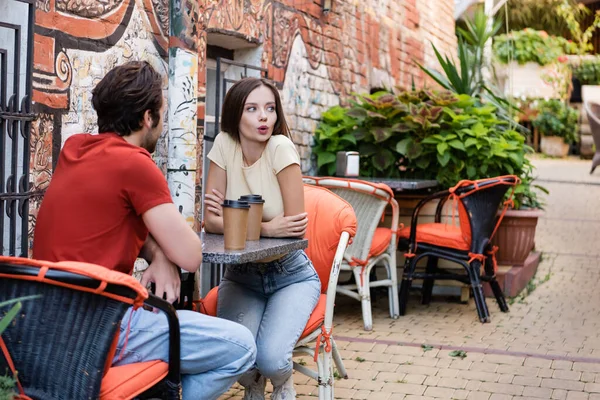 Surprised woman talking near paper cups and boyfriend on cafe terrace — Stock Photo