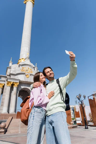 KYIV, UKRAINE - SEPTEMBER 1, 2021: Smiling travelers with backpacks hugging and taking selfie on smartphone on urban street — Stock Photo