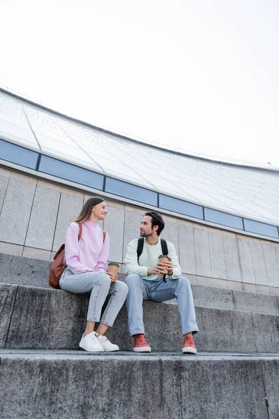 Turistas sonrientes con mochilas y café para ir a hablar cerca del edificio en la calle urbana - foto de stock