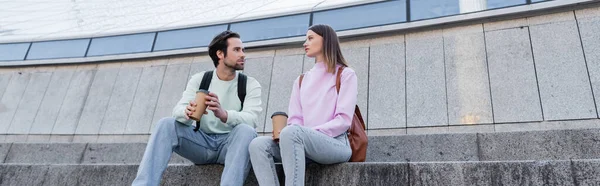 Junge Reisende mit Coffee to go und Rucksäcken reden auf Treppen in der Nähe von Gebäuden in der städtischen Straße, Banner — Stockfoto