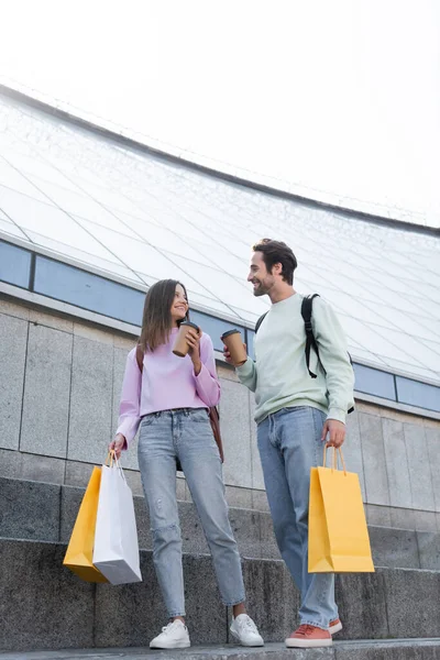 Positive tourists with paper cups and shopping bags walking on urban street — Stock Photo