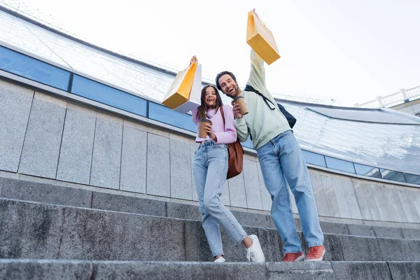 Low angle view of happy travelers with shopping bags and coffee to go looking at camera near building outdoors — Stock Photo