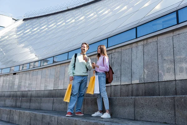 Pareja positiva sosteniendo vasos de papel y bolsas de compras cerca del edificio al aire libre - foto de stock