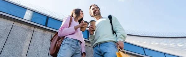 Low angle view of positive couple holding paper cups and shopping bag on urban street, banner — Stock Photo