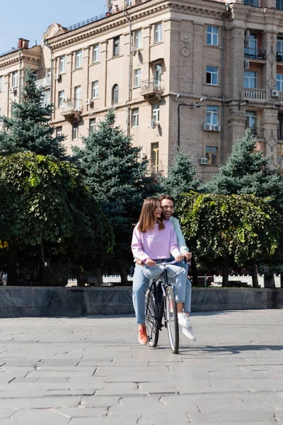 Smiling couple riding bicycle near trees on urban street — Stock Photo