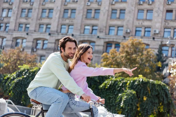 Excited woman pointing with finger while riding bike with boyfriend on urban street — Stock Photo