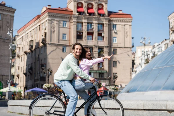 Smiling man riding bicycle with girlfriend pointing with finger on urban street — Stock Photo