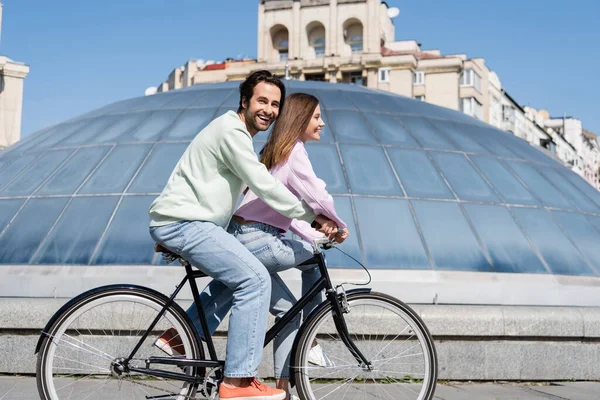 Sorrindo homem olhando para a câmera enquanto pedalava com a namorada na rua urbana — Fotografia de Stock