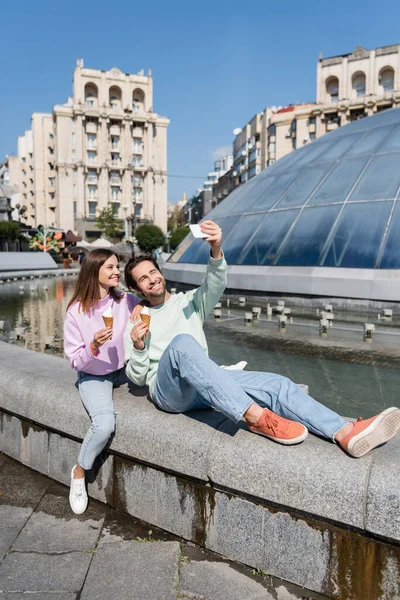 Coppia positiva con gelato scattare selfie su smartphone vicino fontana sulla strada urbana — Foto stock