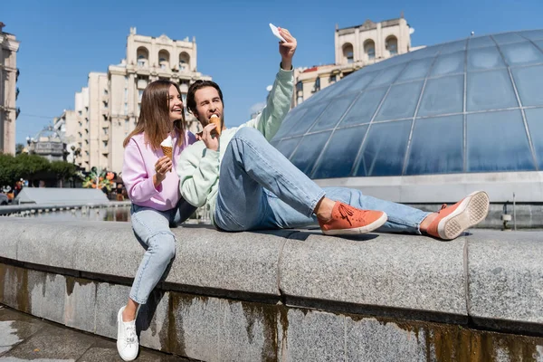 Donna sorridente che tiene il gelato vicino al fidanzato scattare selfie su smartphone sulla strada urbana — Foto stock