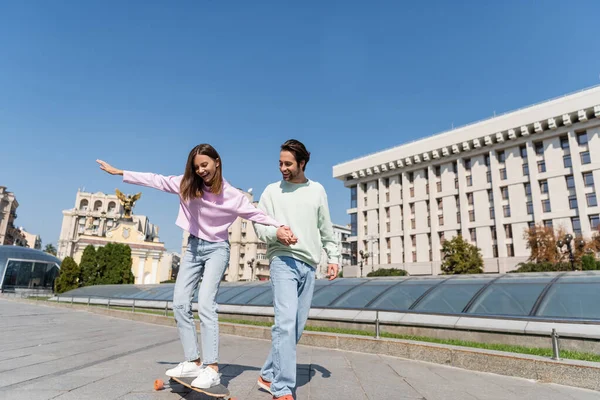 Happy woman riding penny board and holding hand of boyfriend on urban street — Stock Photo