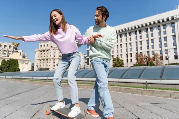 Sonriente hombre cogido de la mano de su novia cabalgando penny board en la calle urbana - foto de stock