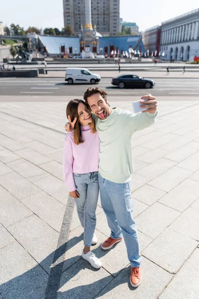 Pareja positiva haciendo gestos y tomando selfie en la calle urbana - foto de stock