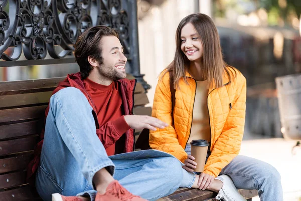 Uomo sorridente che parla con la ragazza con il caffè per andare in panchina sulla strada urbana — Foto stock