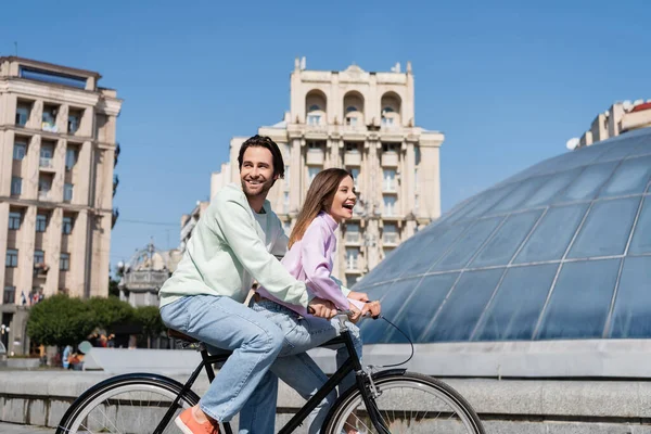 Cheerful couple cycling near building on urban street — Stock Photo