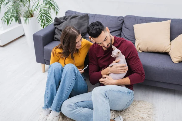 Vista ad alto angolo di donna sorridente che tiene smartphone vicino al fidanzato arabo e al gatto sfinge a casa — Foto stock