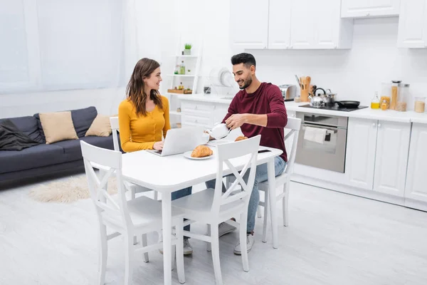 Sorridente coppia multietnica seduta vicino a colazione e laptop in cucina — Foto stock