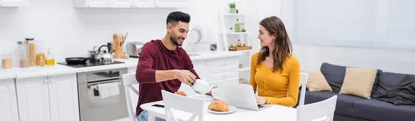 Hombre musulmán positivo verter té cerca de la novia con el ordenador portátil y el desayuno en la mesa en la cocina, pancarta - foto de stock