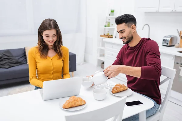 Весела міжрасова пара використовує ноутбук біля сніданку на кухні — стокове фото