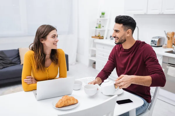 Feliz pareja multiétnica hablando cerca de dispositivos y desayuno en la cocina - foto de stock
