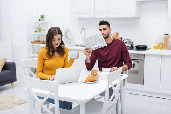 Femme tenant tasse et en utilisant un ordinateur portable près du petit ami arabe avec journal dans la cuisine — Photo de stock