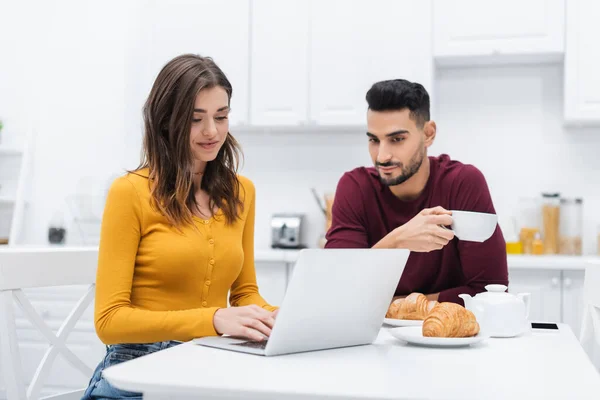 Donna sorridente con computer portatile vicino all'uomo musulmano e colazione in cucina — Foto stock