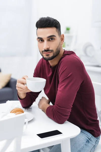 Homem muçulmano segurando copo e olhando para a câmera perto de smartphone e croissant na cozinha — Fotografia de Stock