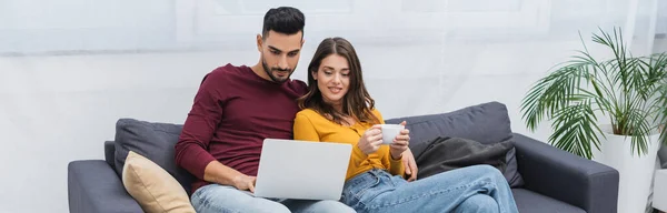 Mujer alegre sosteniendo café cerca novio árabe en el sofá en casa, pancarta - foto de stock