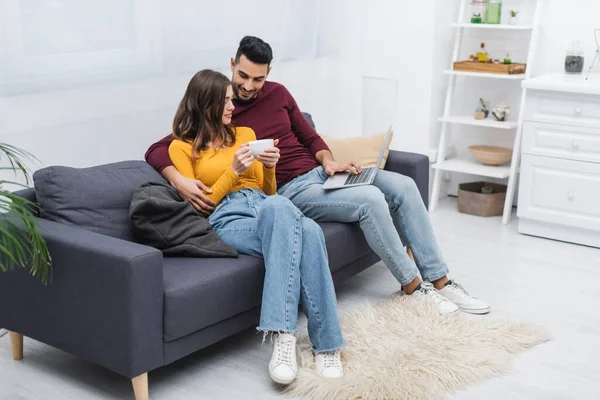 Smiling muslim man hugging girlfriend with cup and using laptop at home - foto de stock