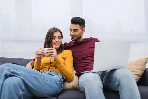 Cheerful woman holding cup of coffee near arabian boyfriend with laptop on couch - foto de stock