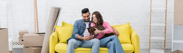Cheerful interracial couple petting bengal cat on couch near packages at home, banner — Stockfoto