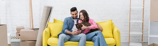 Cheerful interracial couple petting bengal cat on couch near packages at home, banner — Stock Photo