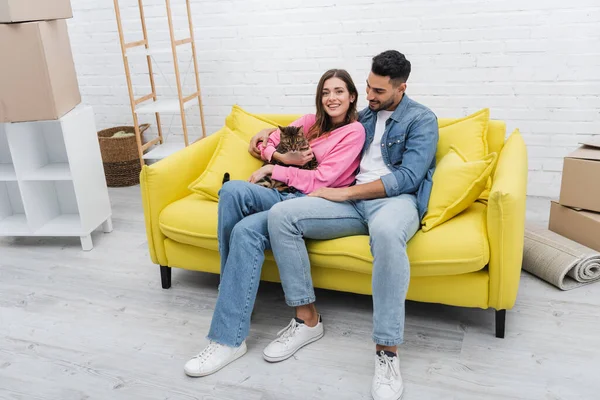 Mujer feliz sonriendo a la cámara mientras sostiene gato bengala cerca de novio musulmán en el sofá - foto de stock