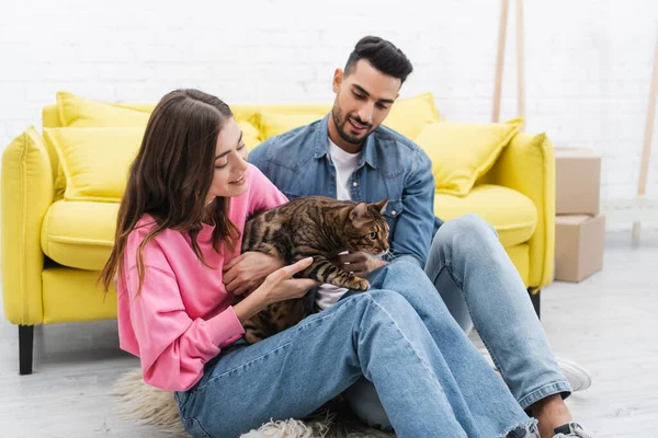 Positive woman holding bengal cat near arabian boyfriend at home — стоковое фото